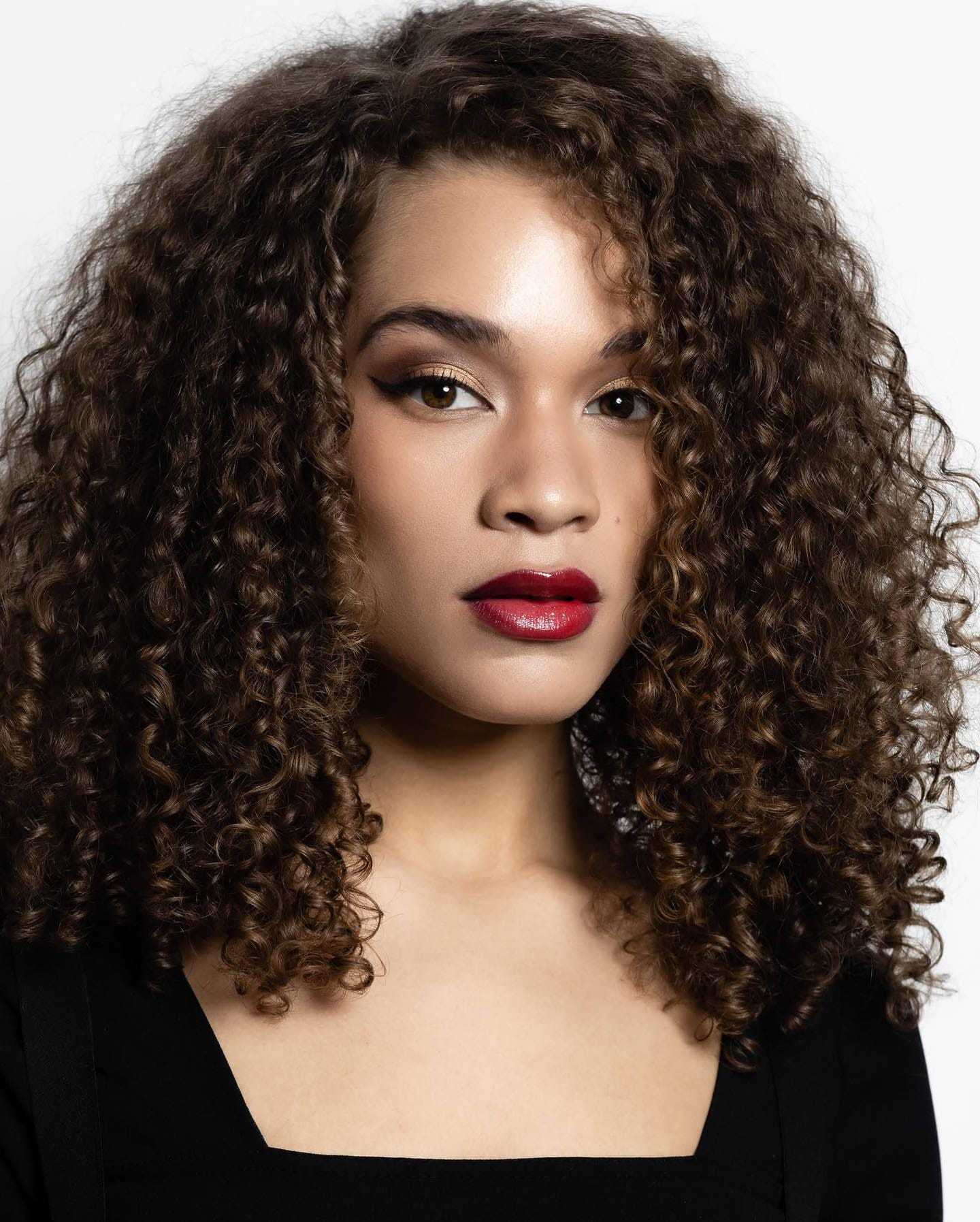 Curly-haired woman with bold makeup gazing confidently against a white background.